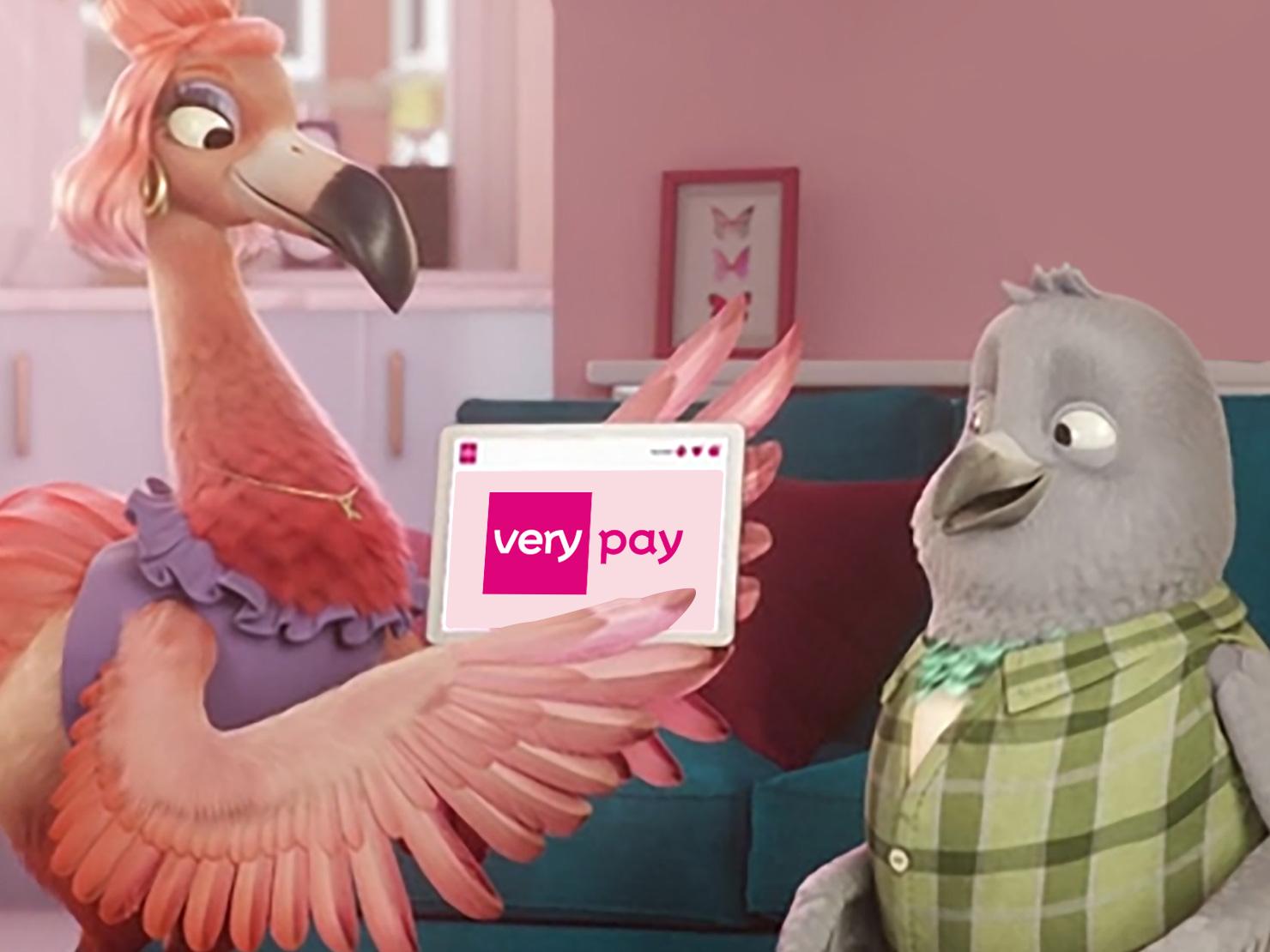 A cartoon flamingo holding a digital tablet displaying the text 'very pay' while engaging with a cartoon grey bird in a colourful living room. The flamingo wears a purple outfit, and the grey bird is in a green plaid shirt.